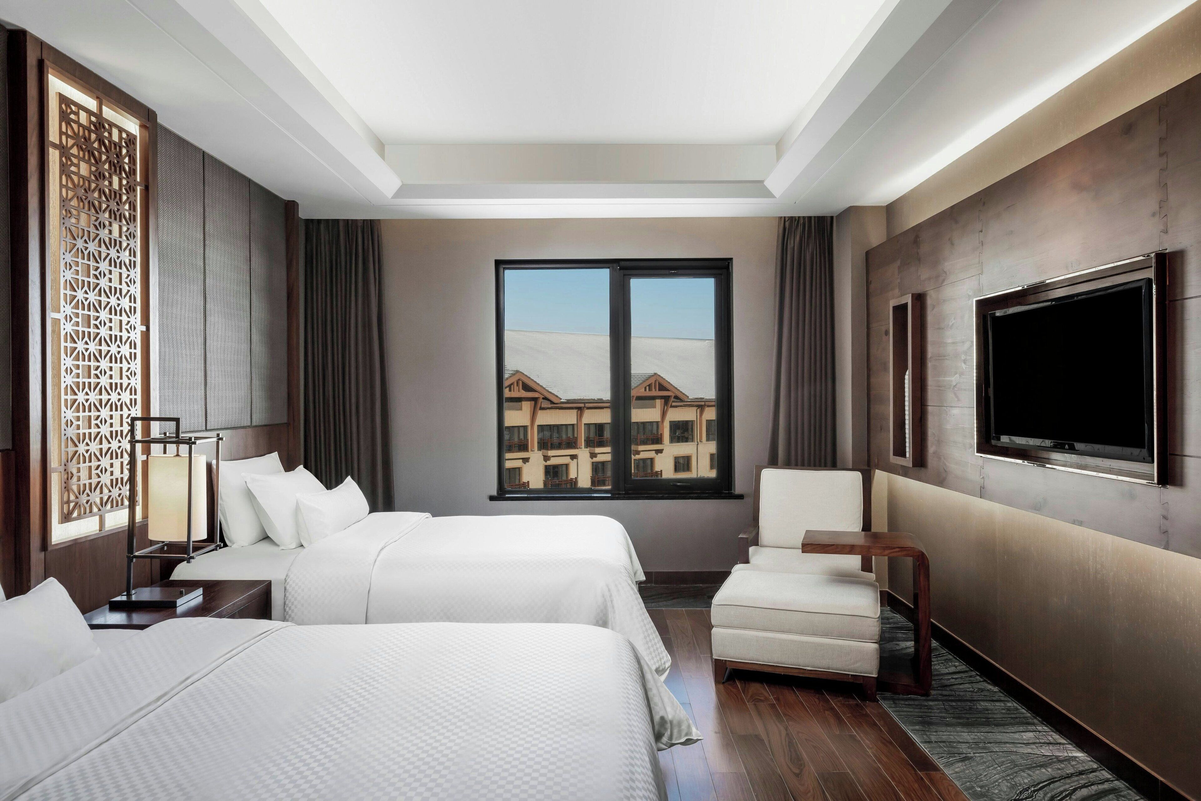 A hotel room with two beds, a TV, a chair, and a large window with a view of buildings.