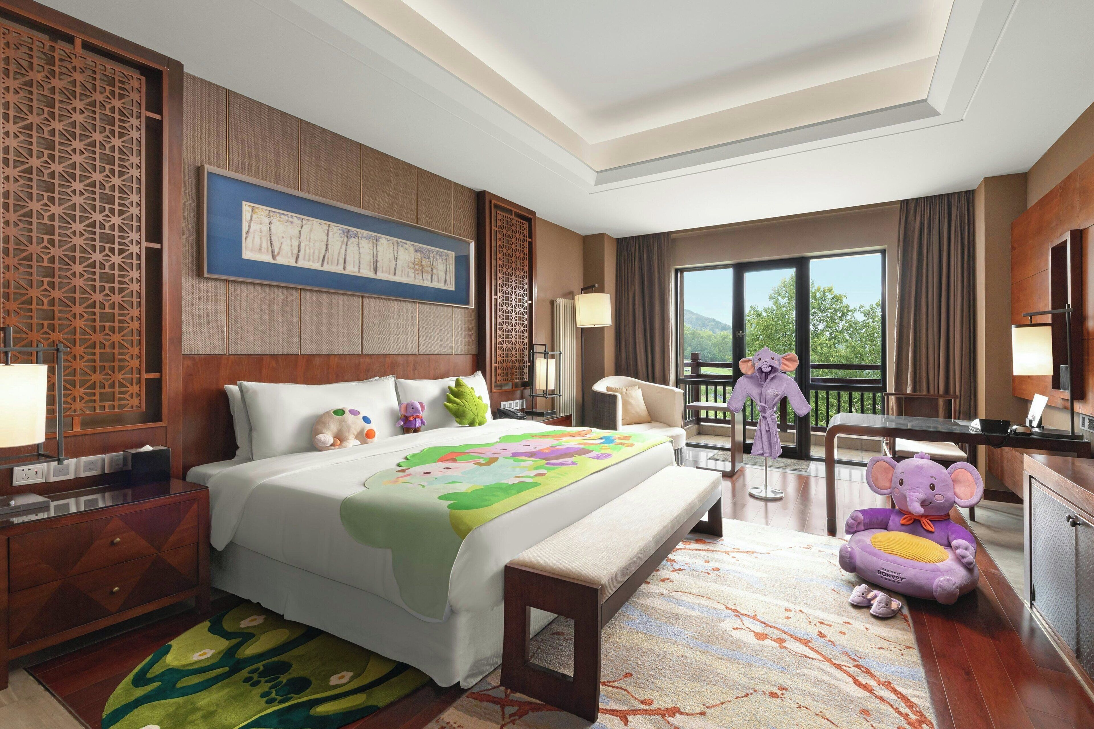 A hotel room with a large bed, a bench, a dresser, a nightstand, a lamp, a window with a view of greenery, and a decorative screen.