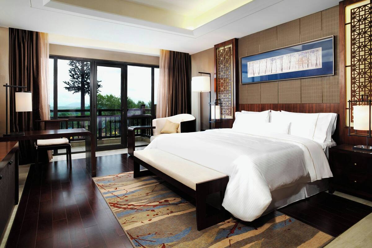 A hotel room with a large bed, a desk, a chair, and a balcony with a view of trees.