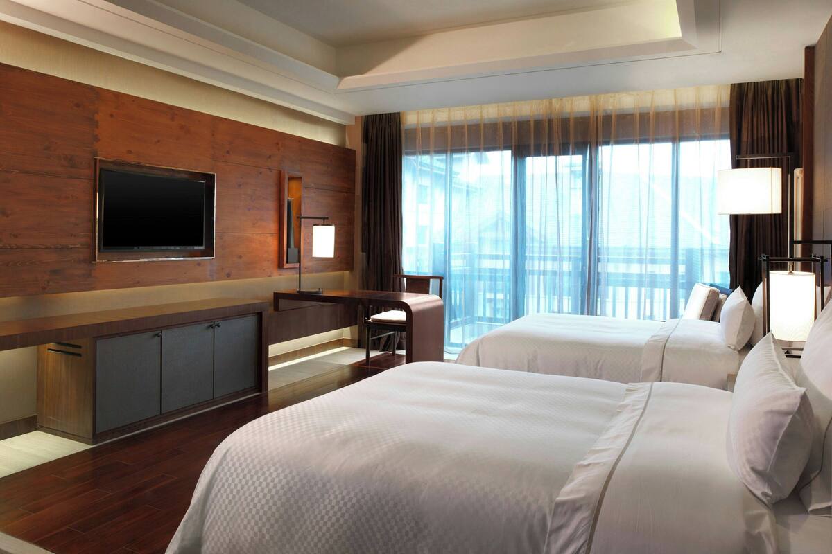 A hotel room with two beds, a TV mounted on the wall, a desk with a chair, and a large window with curtains.