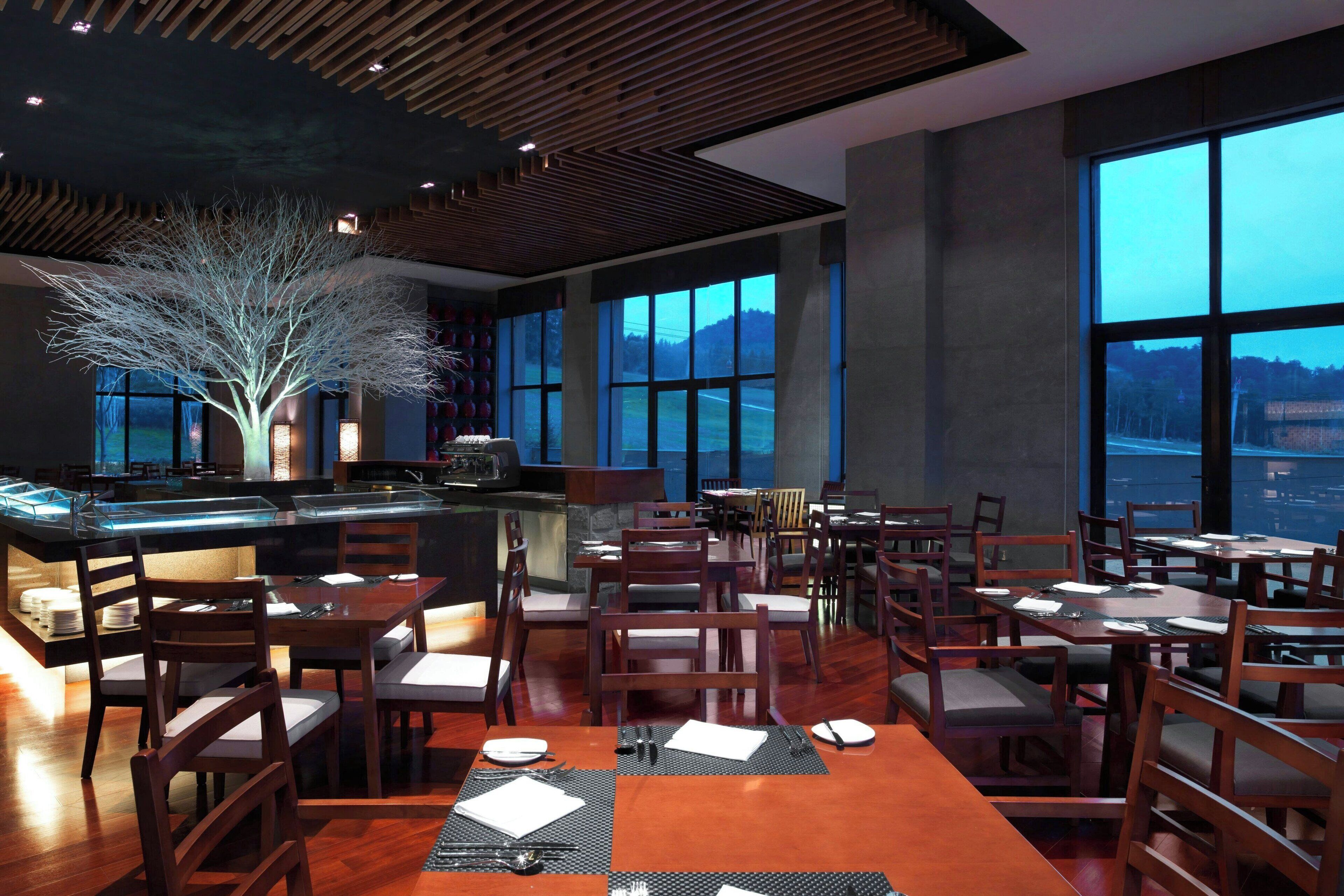 A restaurant with wooden furniture, large windows, and a tree sculpture.
