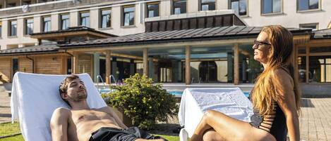 Un hombre y una mujer descansando en tumbonas junto a una piscina, con un edificio con balcones al fondo.