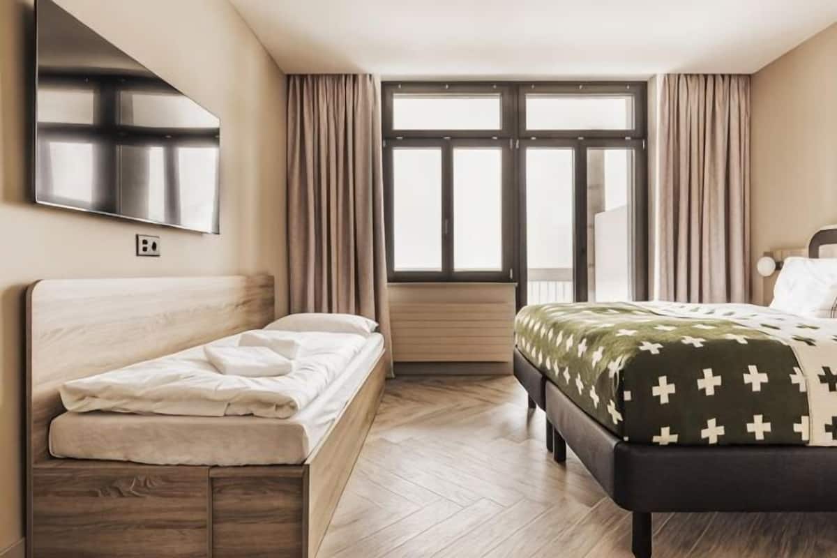 A hotel room with a bed, a TV, a window with curtains, and a wooden floor.