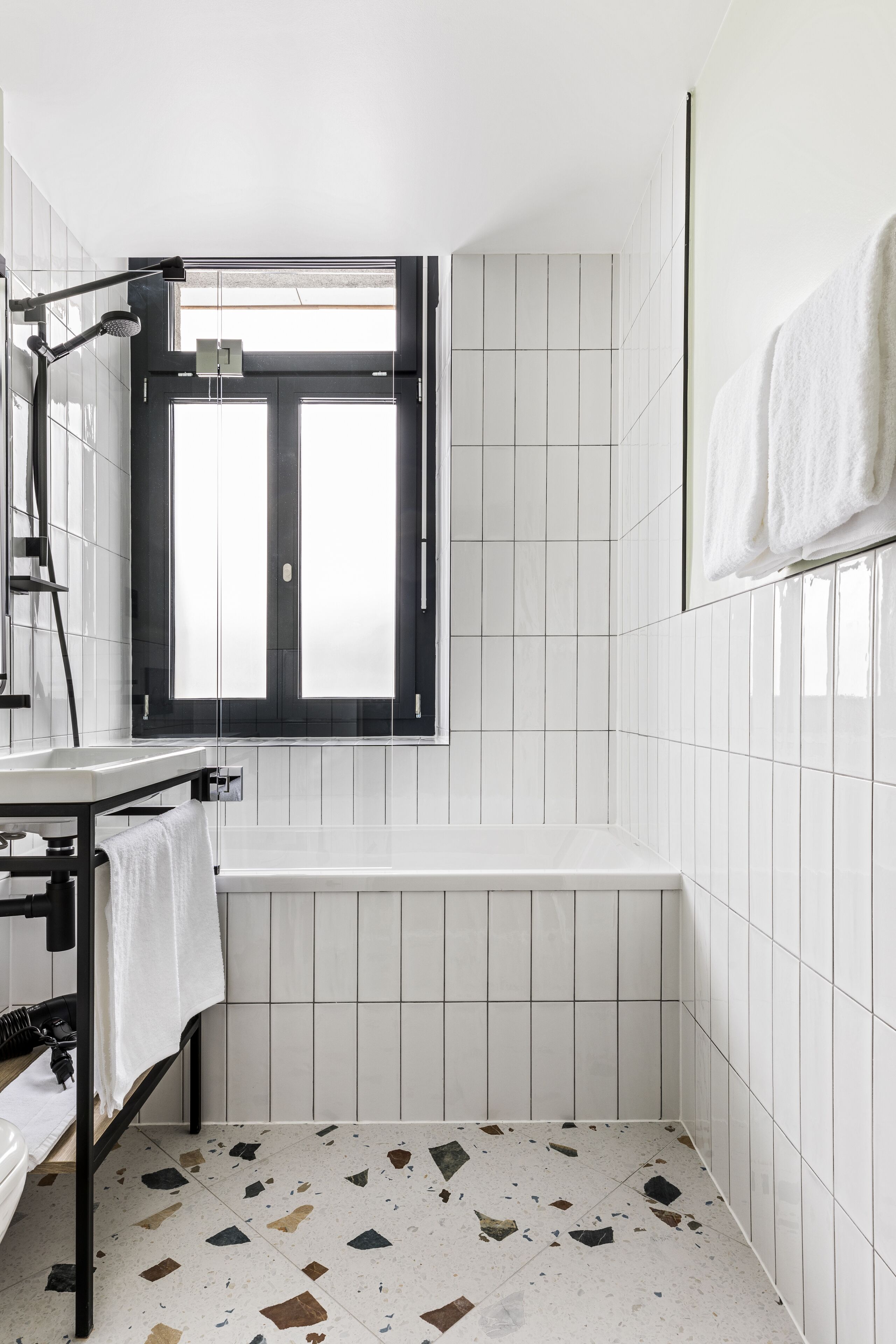 A modern bathroom with white tiles, a bathtub, a shower with a handheld showerhead, and a window with black shutters.