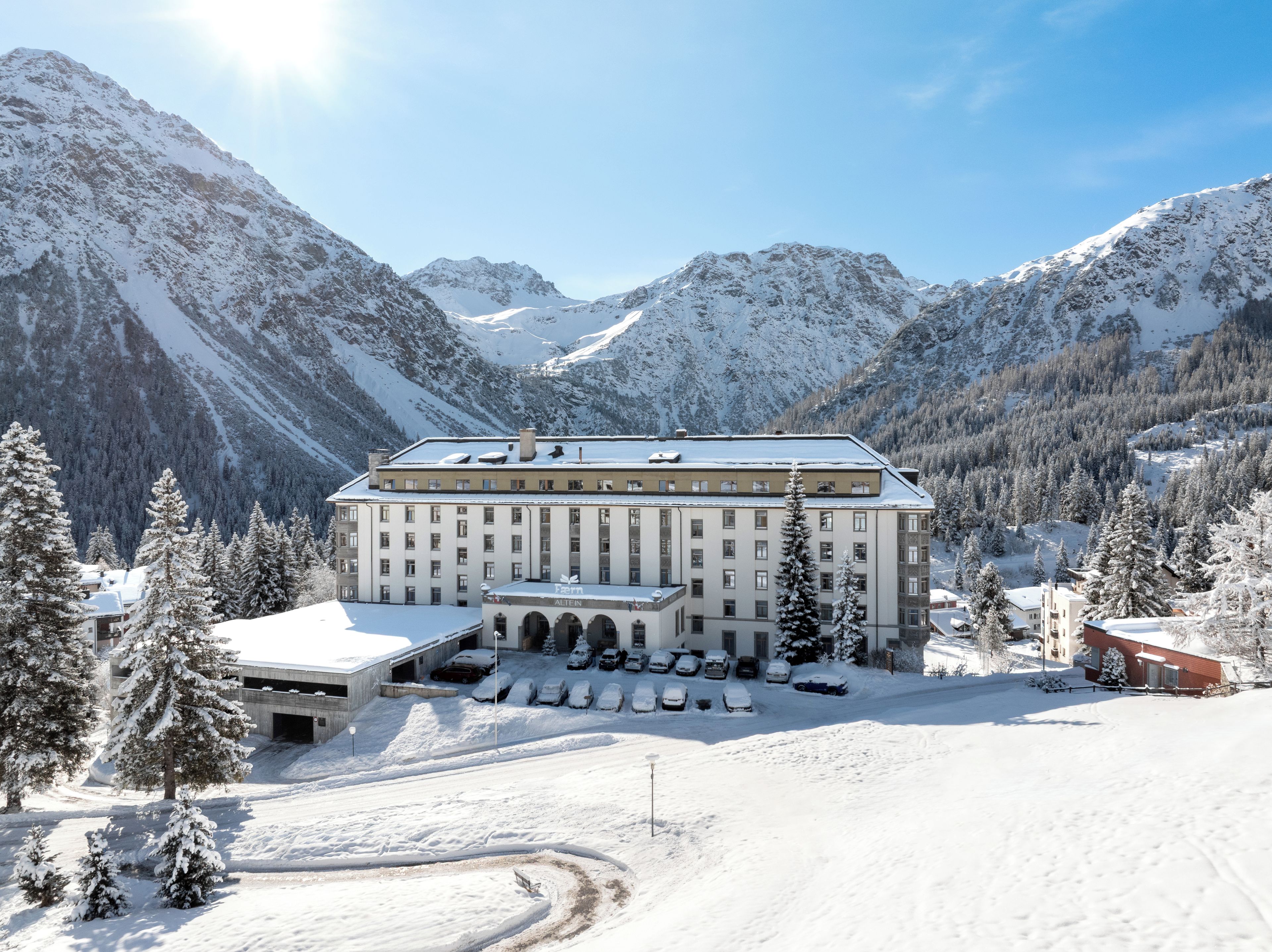 A large building with multiple windows and a flat roof, surrounded by snow-covered mountains and trees.