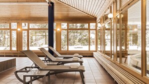 Un espacio interior luminoso con suelo de madera y ventanas amplias, que presenta sillones de sol y vistas a árboles nevados al exterior.