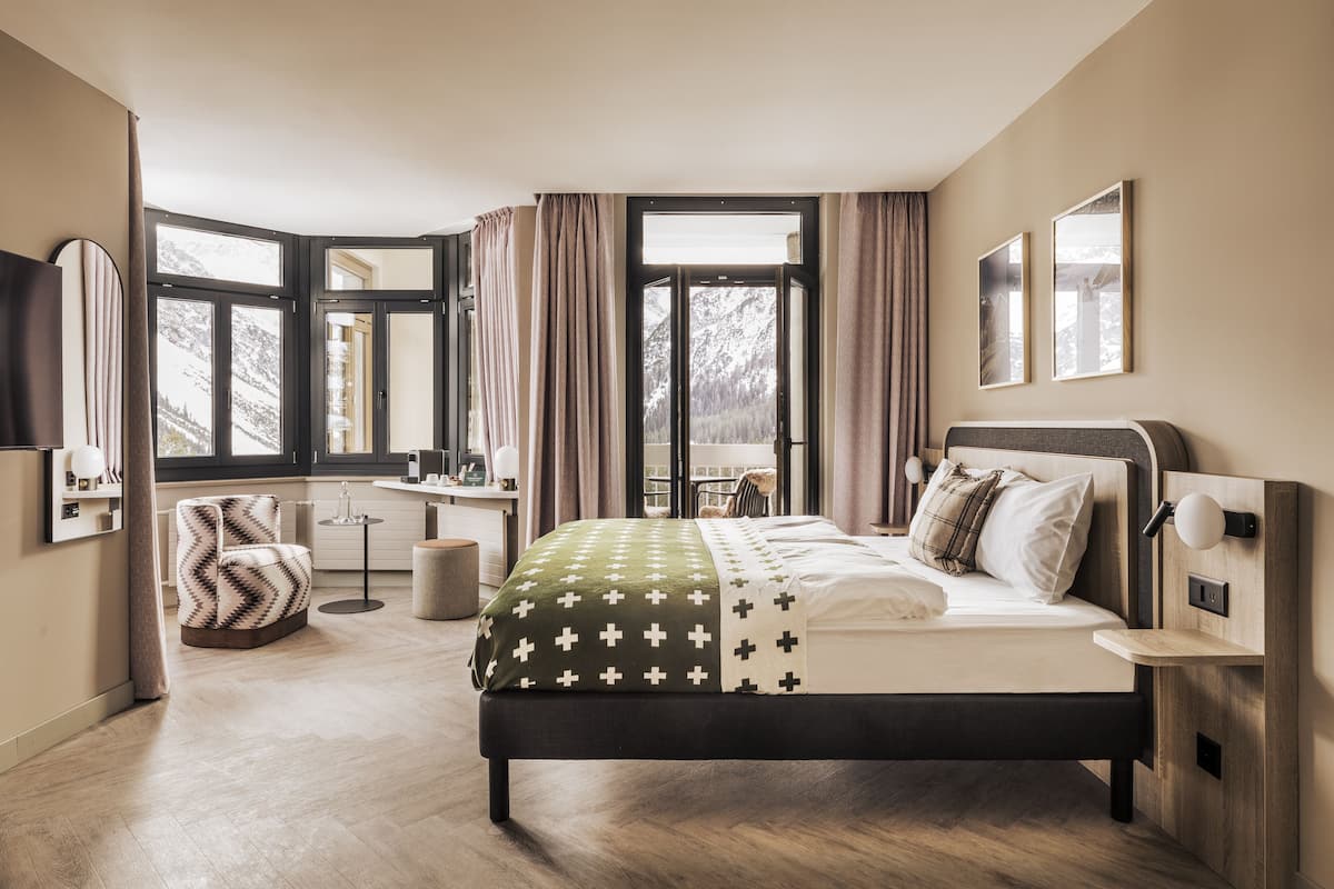 A modern bedroom with a large bed, a sitting area with a chair, a small table, and a view of mountains through large windows.