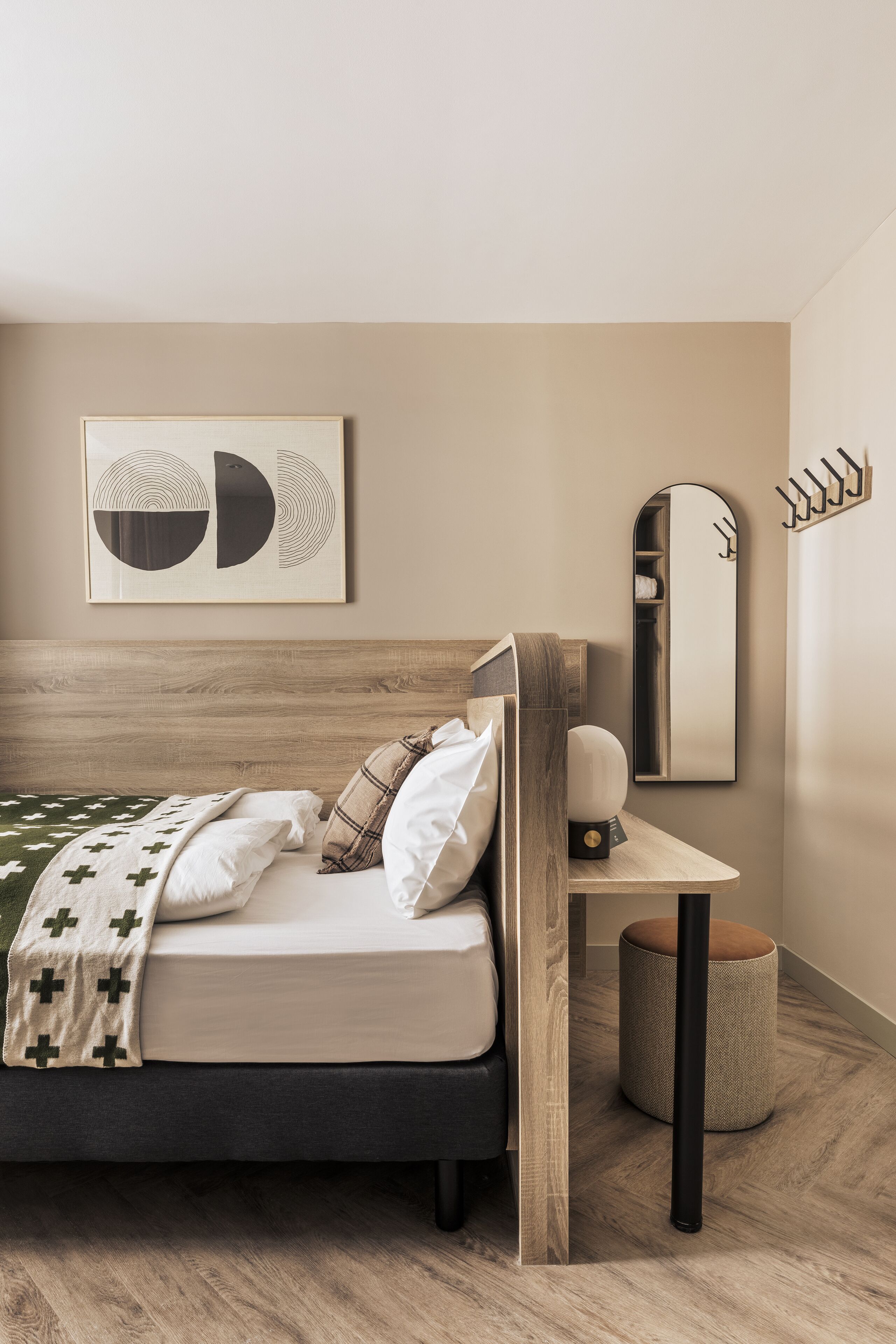 A modern bedroom with a wooden headboard, a bed with a green and white blanket, a round stool, a bedside table with a lamp, and a wall-mounted mirror.