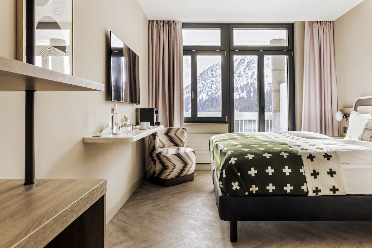 A modern hotel room with a bed, a desk, a chair, and a large window with a mountain view.