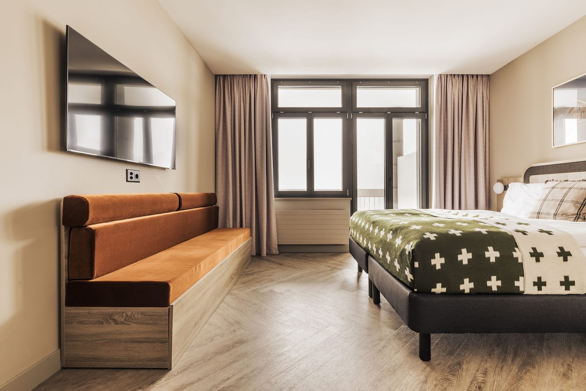 A modern bedroom with a sofa, a bed with a patterned bedspread, a large window with curtains, and a mounted TV.