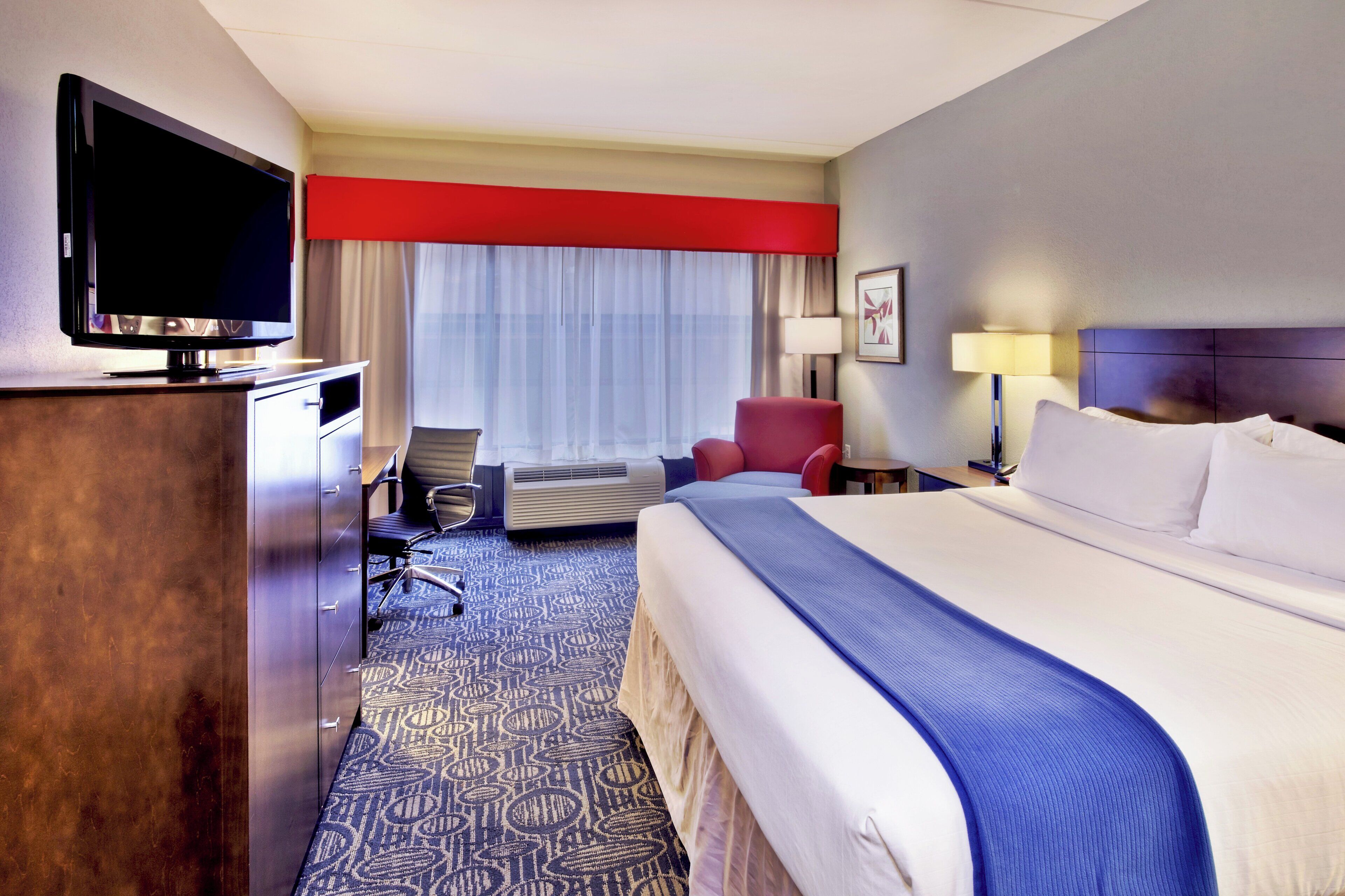 Une chambre d’hôtel avec un lit, un bureau équipé d’un ordinateur, une chaise, un téléviseur et une fenêtre avec des rideaux.
