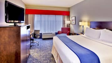 Habitación de hotel con una cama grande, un escritorio con televisor de pantalla plana, un sillón rojo, una lámpara de escritorio y una ventana con cortinas traslúcidas.