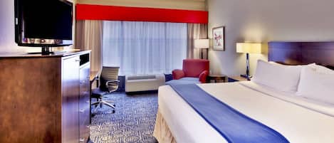 Habitación de hotel con una cama grande, un escritorio con televisor de pantalla plana, un sillón rojo, una lámpara de escritorio y una ventana con cortinas traslúcidas.