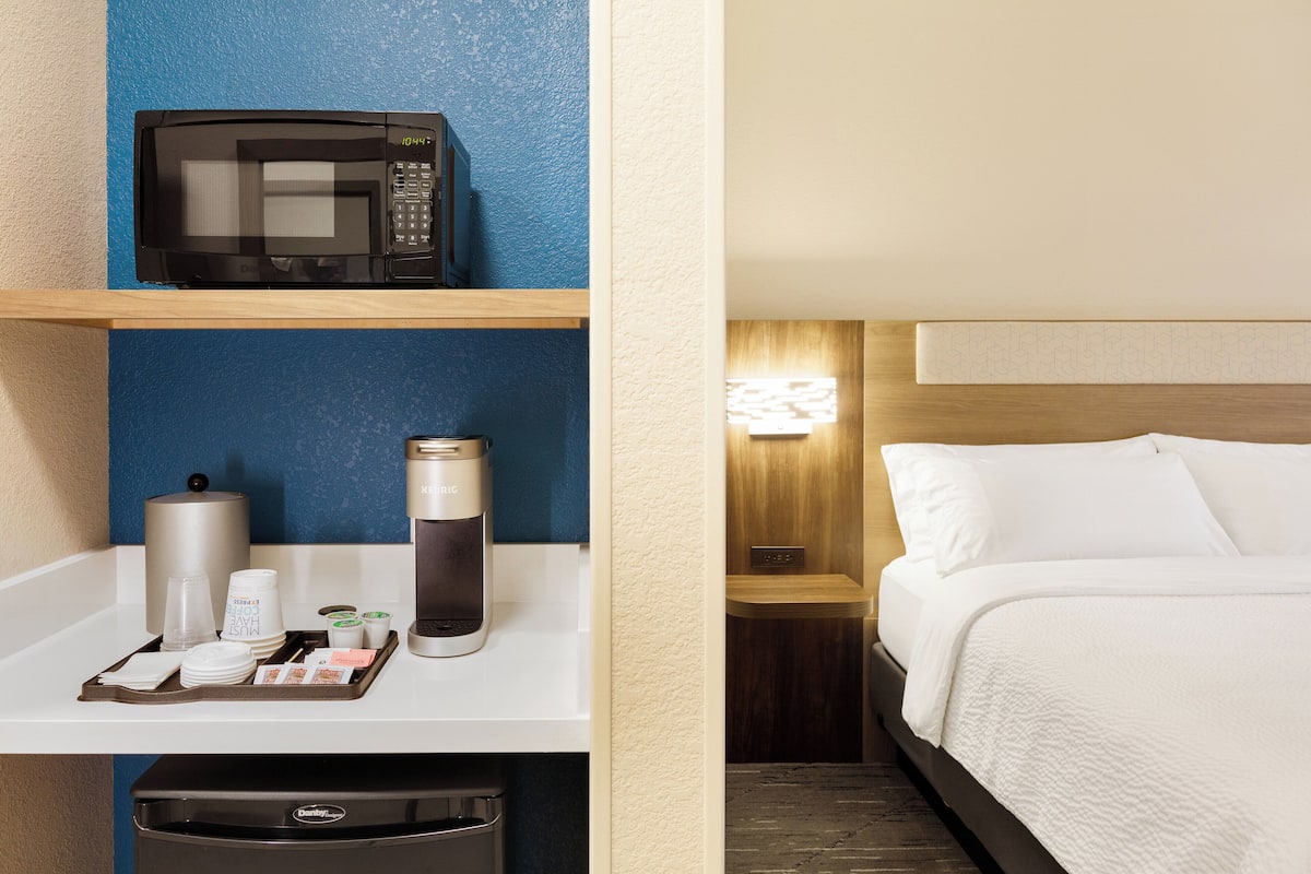 A hotel room with a microwave on a shelf, a coffee maker, and a trash bin.