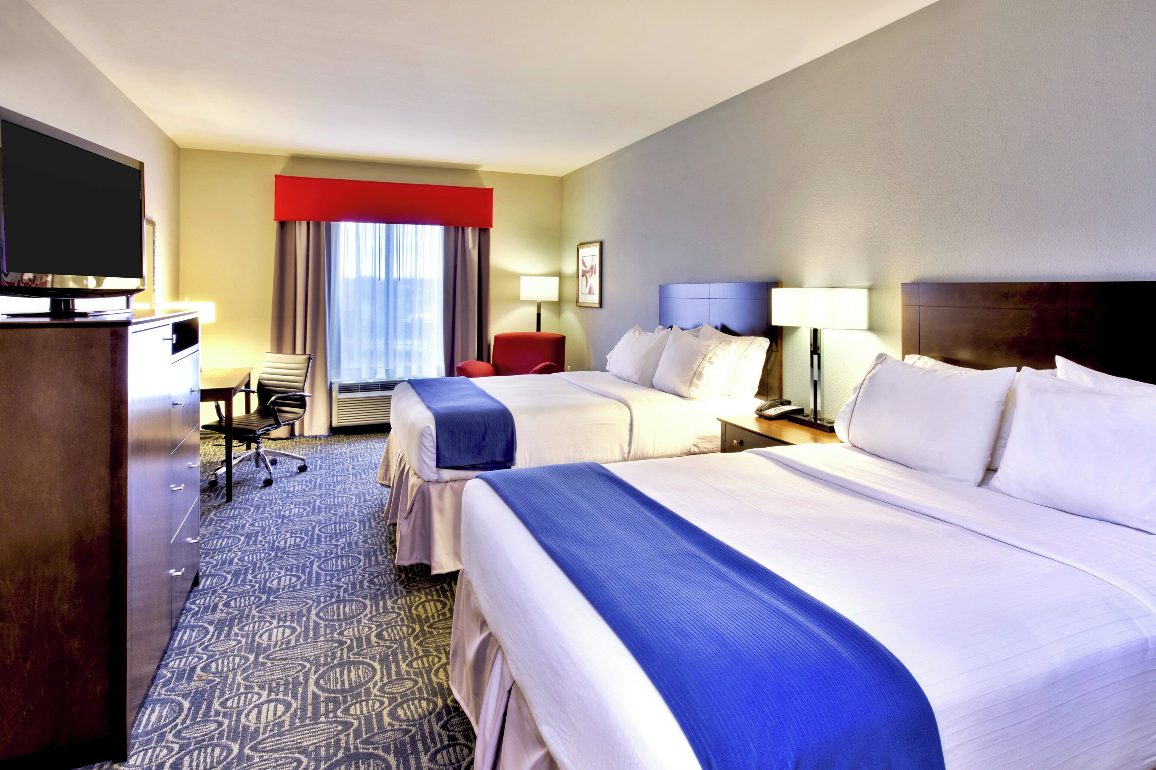 A hotel room with two beds, a desk, a television, and a window with curtains.