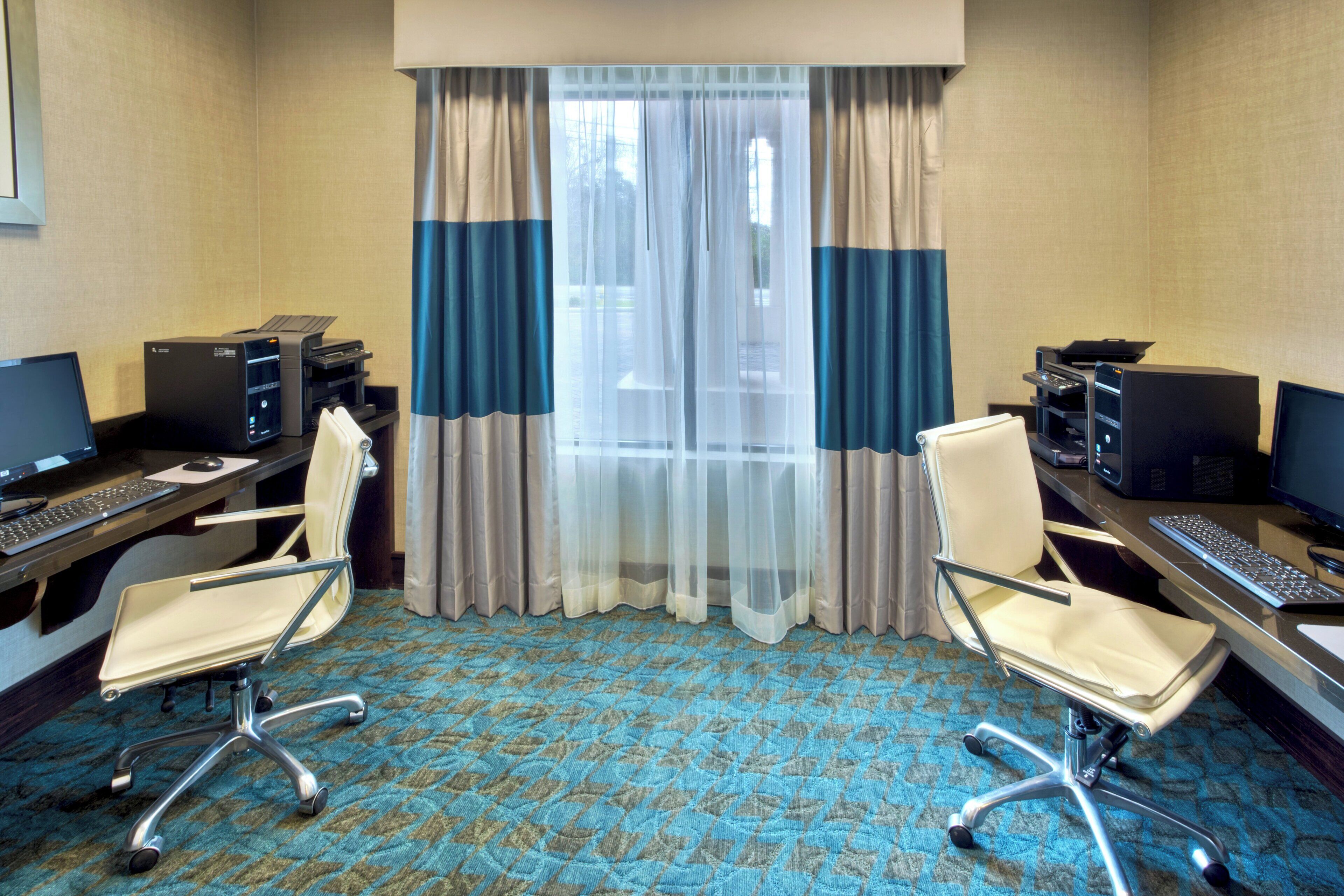 Une chambre d’hôtel  équipée de deux bureaux, chacun comprenant un ordinateur et un clavier, une chaise et une grande fenêtre avec des rideaux légers.
