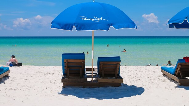 Een strand met twee blauwe parasols met de tekst 'Pawny Jack', houten ligstoelen en een helder uitzicht op de oceaan.