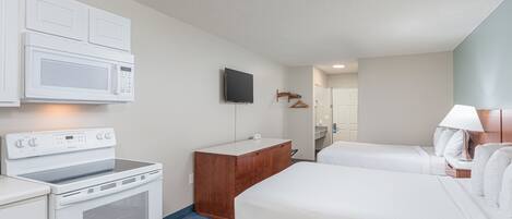 A hotel room with a kitchenette, a TV, a wooden dresser, two beds, and a lamp.