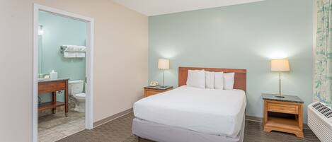 Habitación de hotel con cama, dos mesitas de noche con lámparas, una mesita auxiliar de madera, un baño con inodoro y lavabo, y una puerta que da al baño.