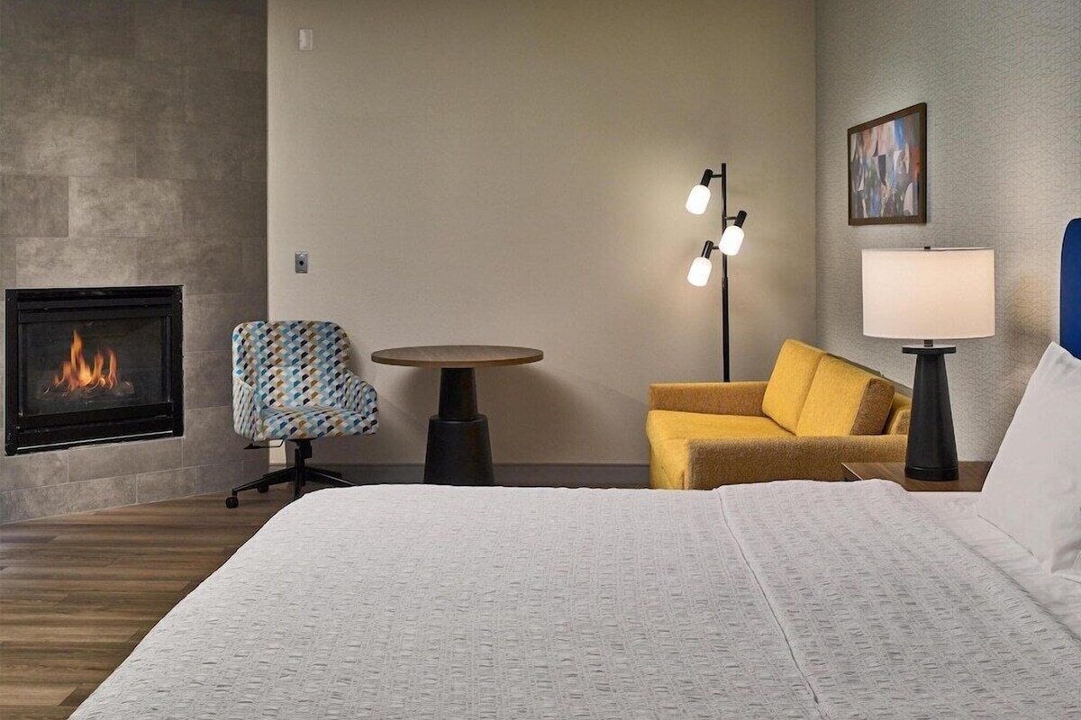 A hotel room with a bed, a sofa, a chair, a table, a lamp, a painting, and a fireplace.