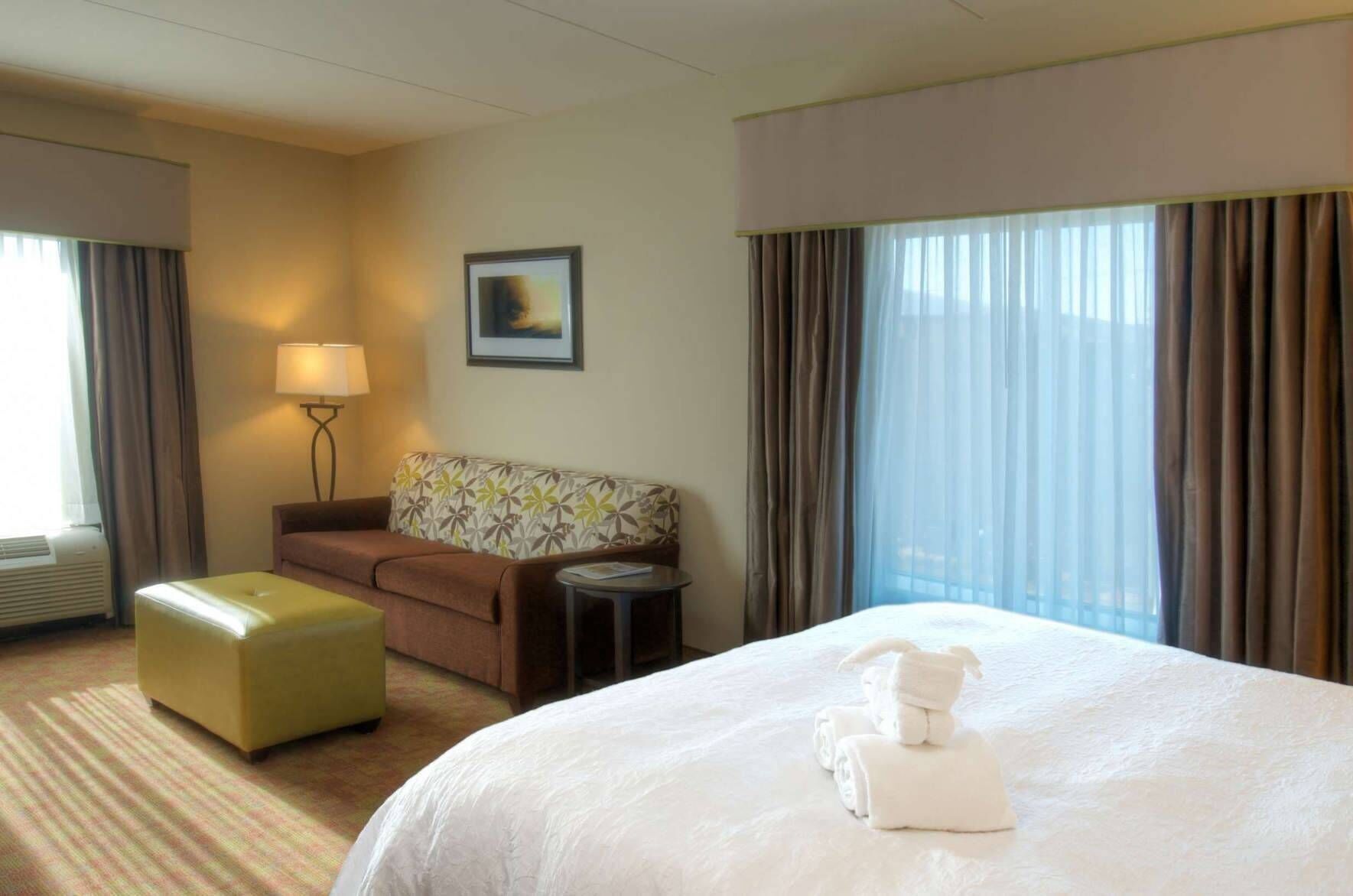 A hotel room with a bed, a sofa, a small table, a lamp, and a window with curtains.