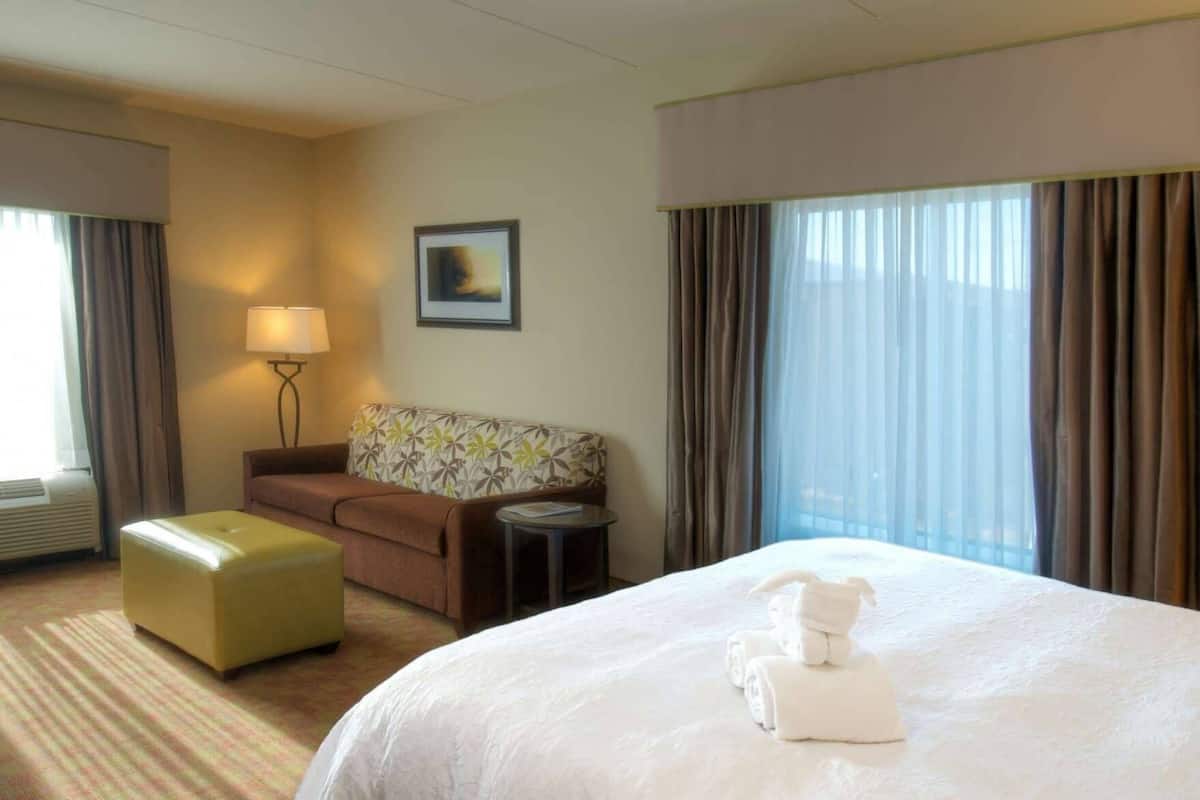 A hotel room with a bed, a sofa, a small table, a lamp, and a window with curtains.