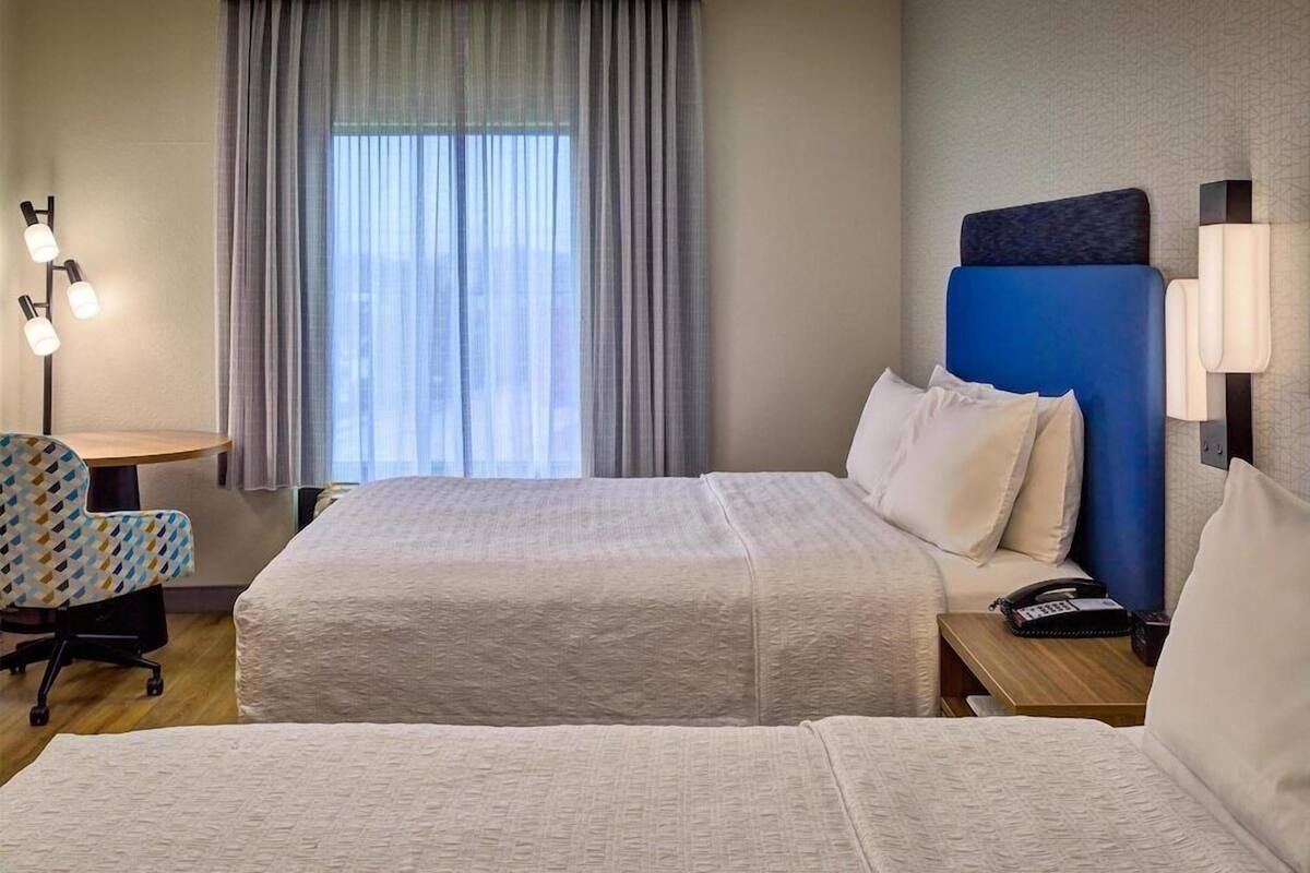 A hotel room with two beds, a desk, a chair, and a large window with curtains.
