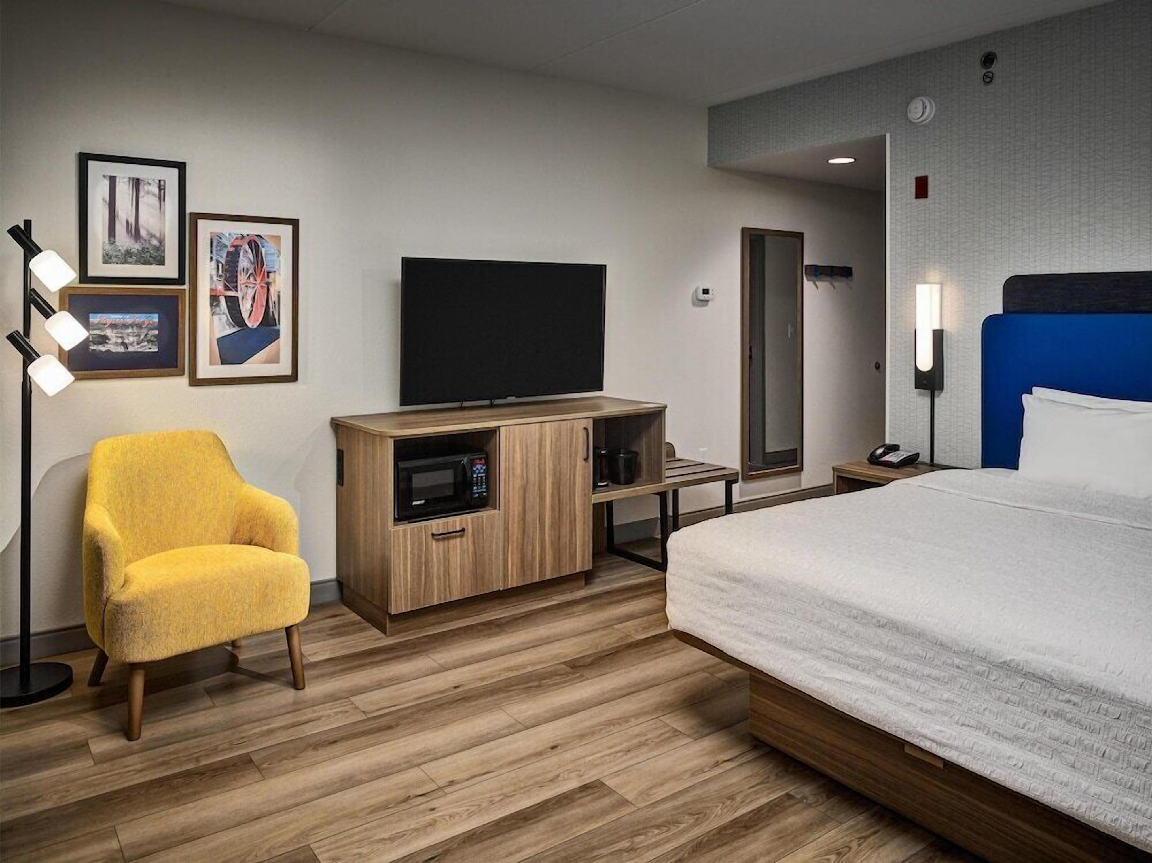 Een hotelkamer met een bed, een gele fauteuil, een televisie, een magnetron, een spiegel en een nachtkastje.