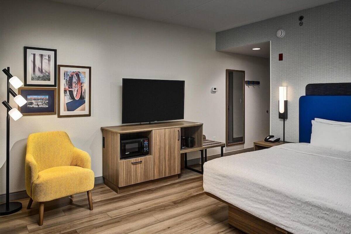 A hotel room with a bed, a yellow armchair, a TV, a microwave, a mirror, and a bedside table.