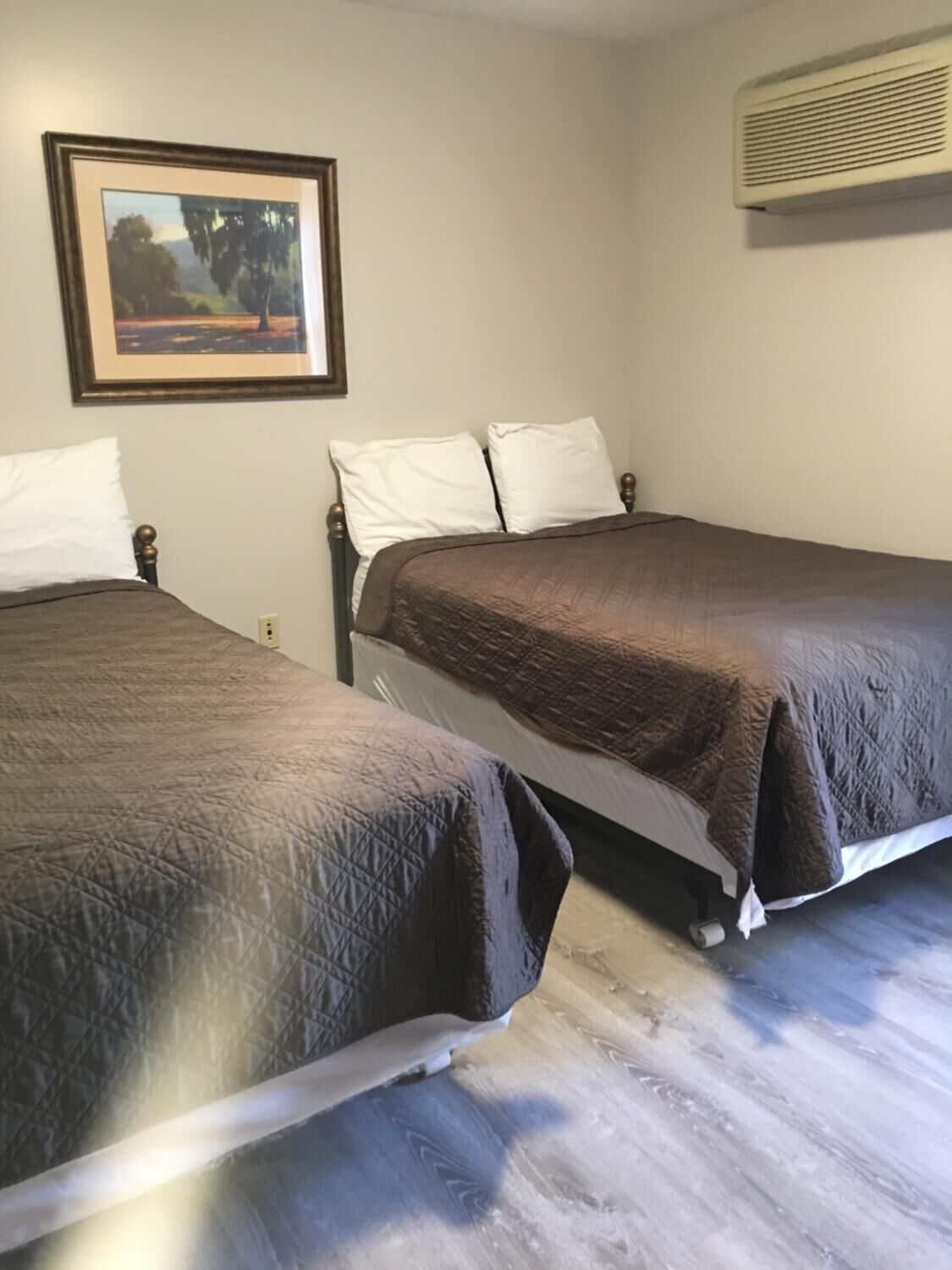 Duas camas de solteiro com roupa de cama marrom, um quadro de paisagem na parede e um ar-condicionado fixado na parede.