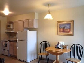 A compact kitchen with a refrigerator, stove, microwave, and wooden cabinets. There is a dining table with two chairs and a hanging light fixture. - All Suites Inn Budget Host (Lewisburg)