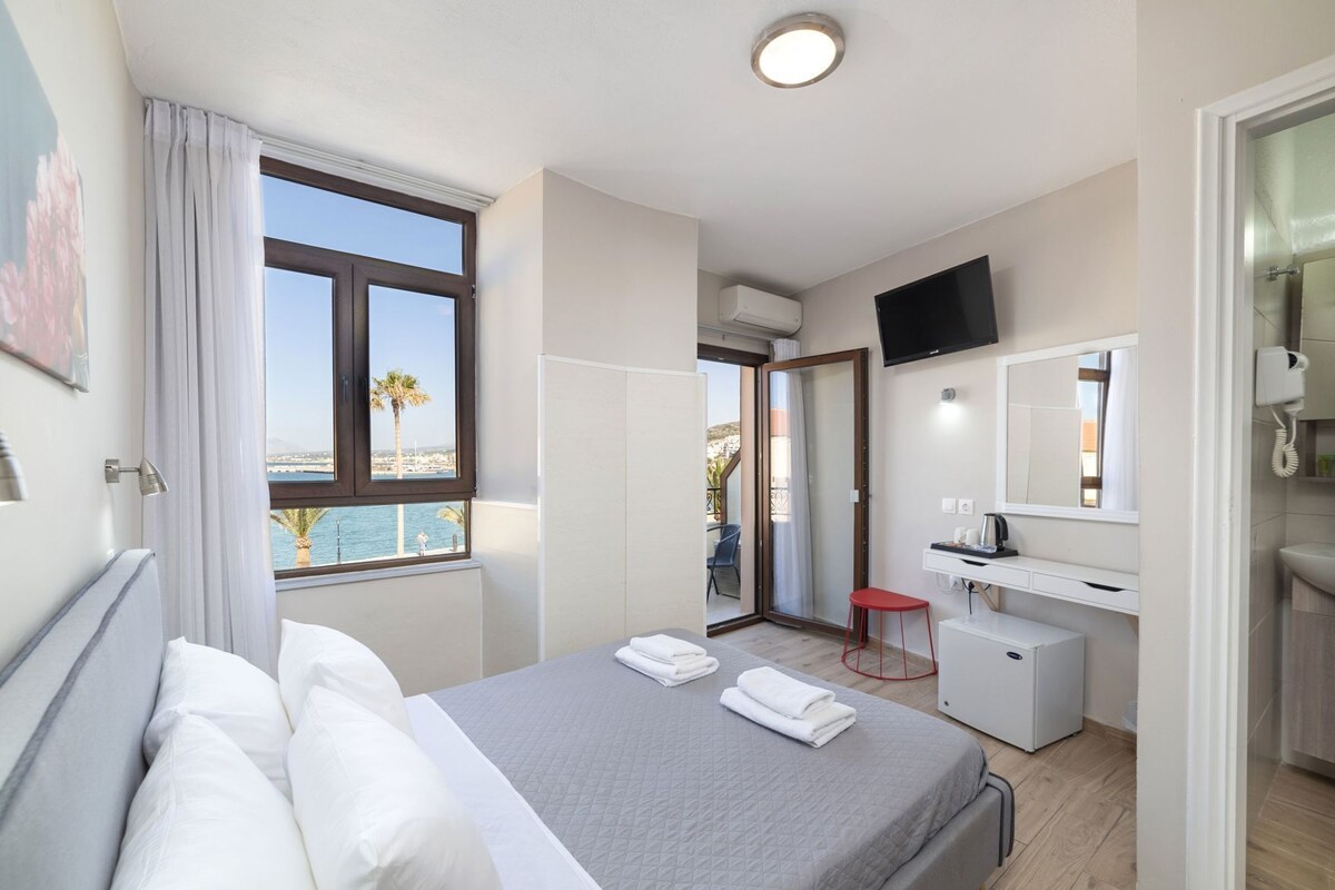 A hotel room with a bed, a desk, a TV, and a view of the sea.
