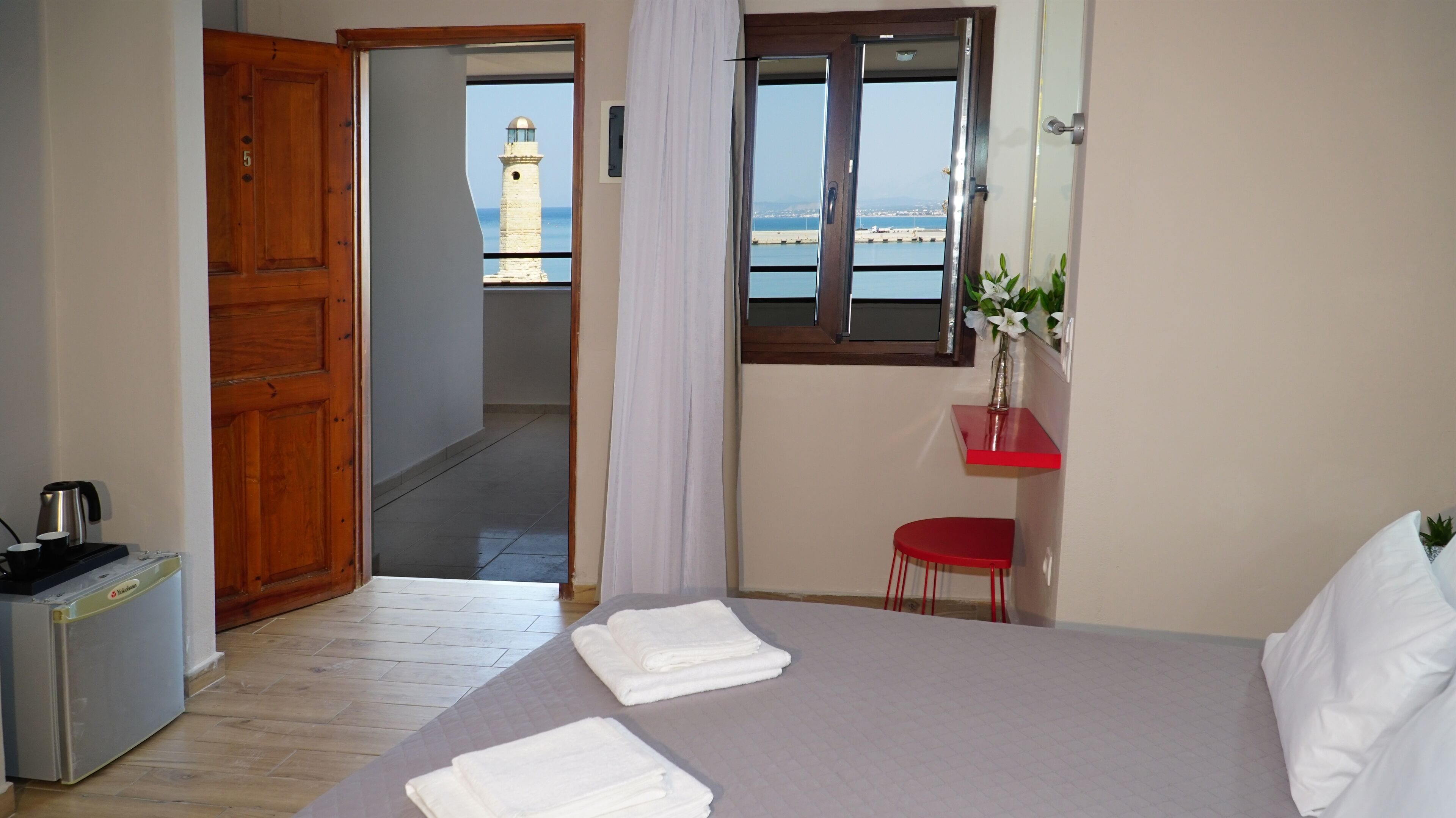Ein Schlafzimmer mit einem Bett, einem roten Sessel, einem kleinen Tisch mit einer Vase und Blick auf einen Leuchtturm durch ein Fenster.