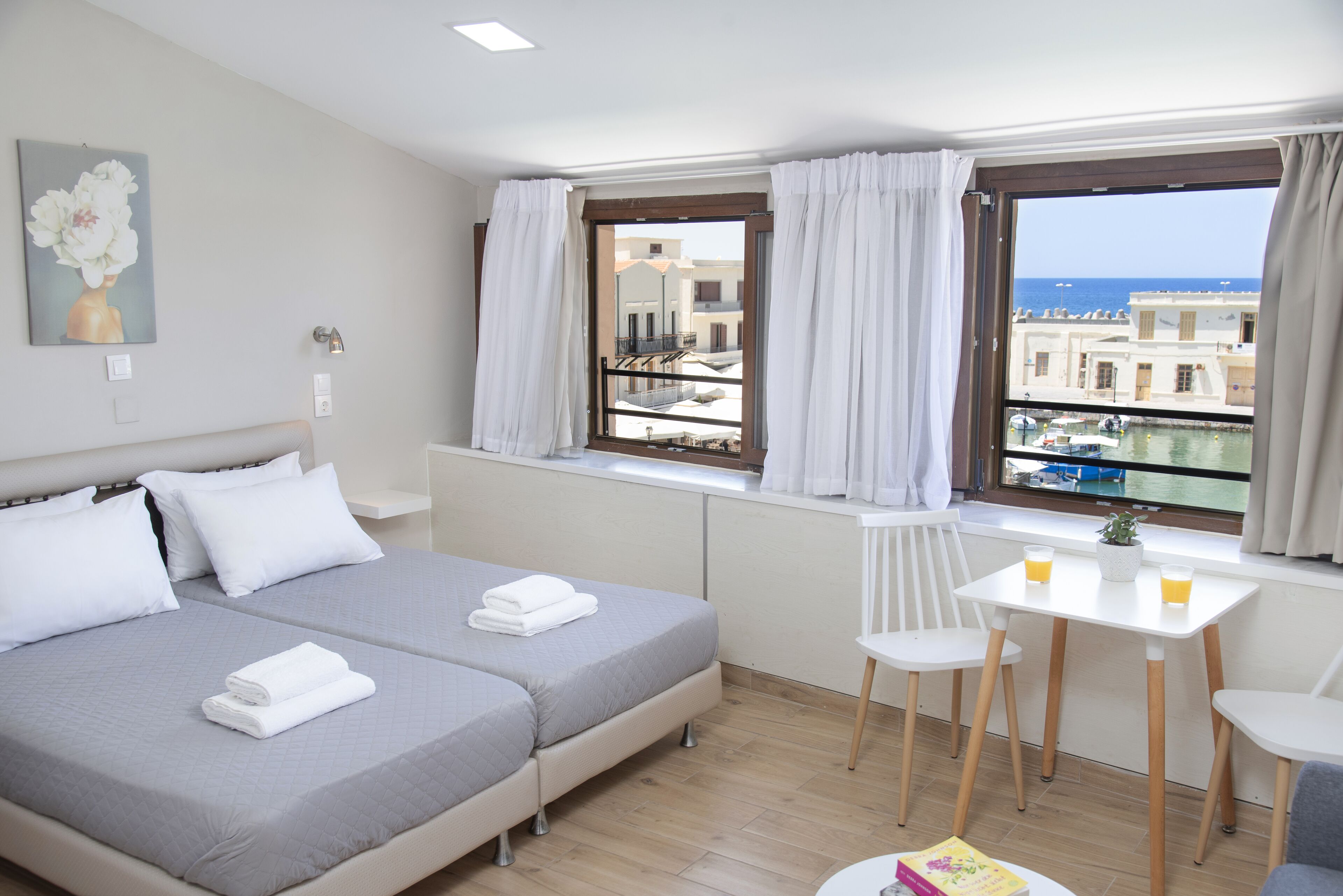 Hotelkamer met twee bedden, een klein tafeltje met stoelen, een raam met gordijnen en uitzicht op gebouwen en water.