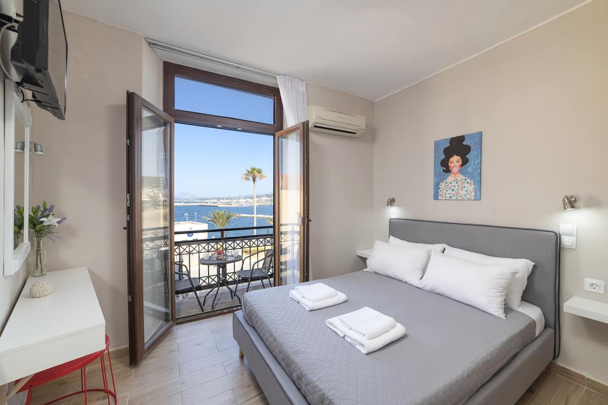 A bedroom with a bed, a desk, a chair, and a balcony with a view of the sea.
