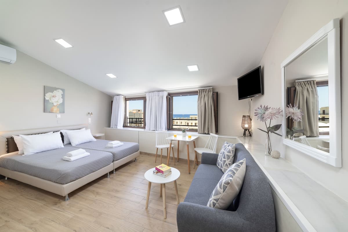 A modern, well-lit room with a bed, sofa, and a small table. There are windows with curtains, a flat-screen TV, and a view of the sea.