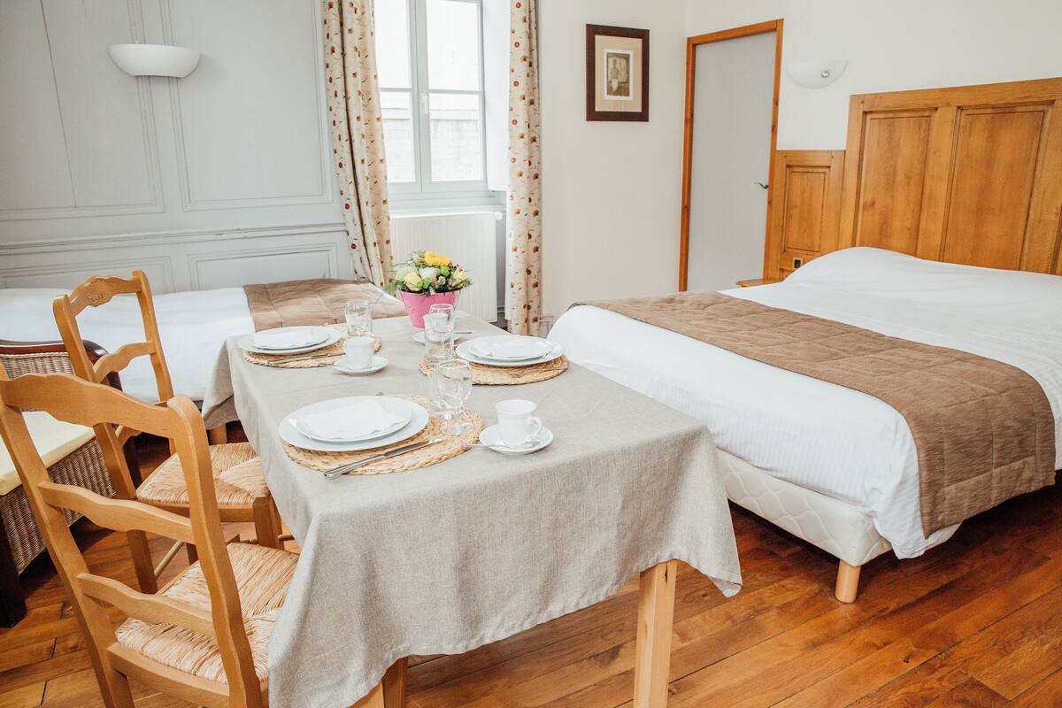 Una habitación ordenada con una cama, una mesa de comedor para dos personas y un ventanal con cortinas.