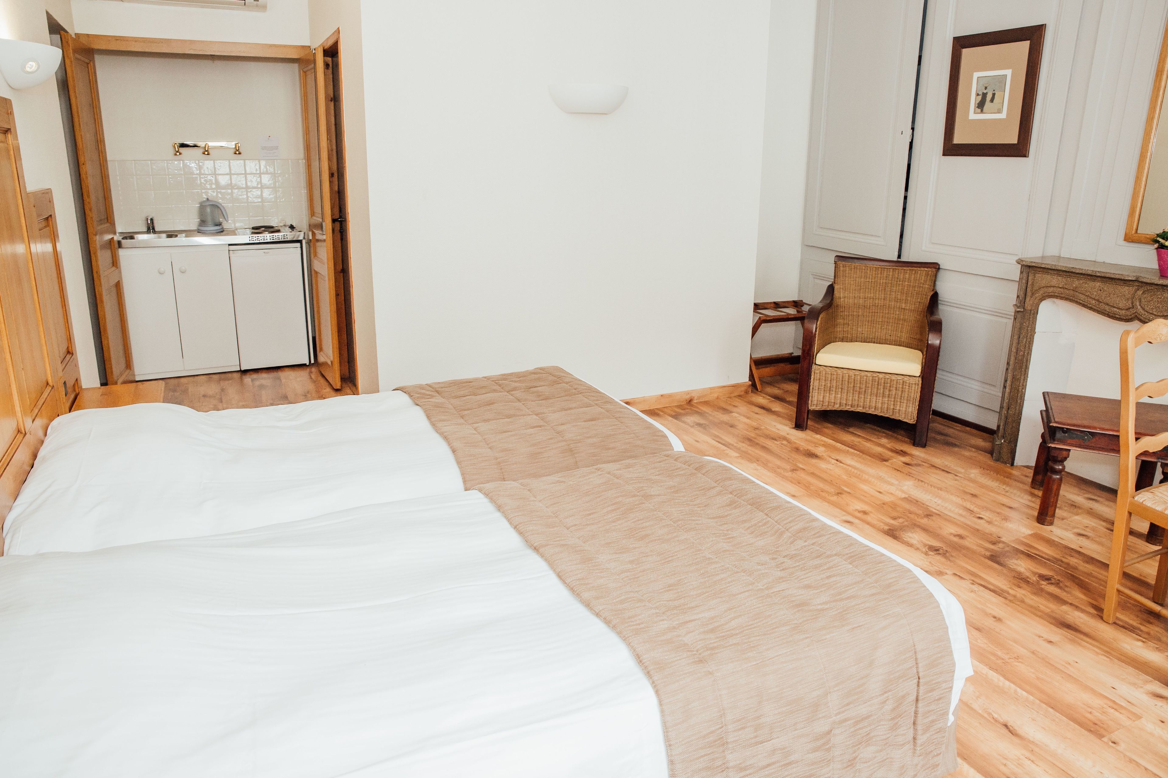 A hotel room with a bed, a chair, a small table, and a kitchenette in the background.