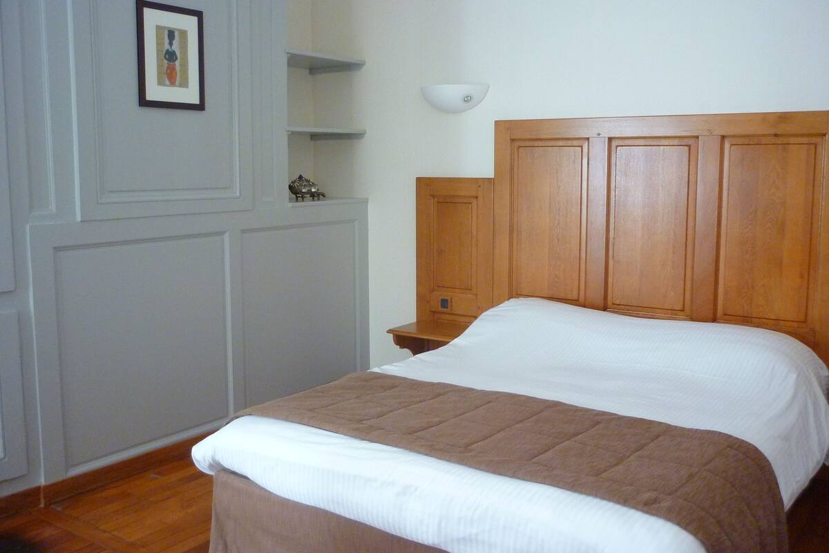 Una cama bien hecha con cabecera de madera, una mesita de noche y un estante de pared con un cuadro enmarcado.