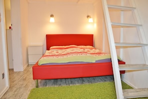 A bedroom with a red bed, two bedside lamps, a green rug, and a white ladder.