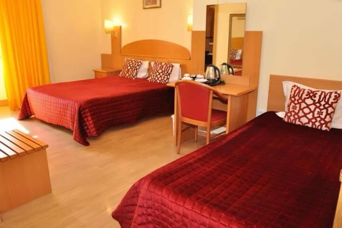 Quarto de hotel com duas camas de solteiro, cada uma com colcha vermelha e almofadas combinando. Há uma escrivaninha de madeira com uma cadeira, um espelho e uma mesinha com chaleira e chávenas.