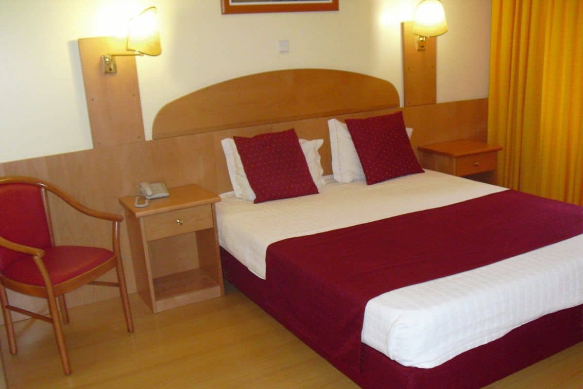 Quarto de hotel com cabeceira de madeira, uma cama de solteiro com roupa de cama vermelha e branca, mesa de cabeceira de madeira com abajur, uma cadeira vermelha e uma escrivaninha de madeira com telefone.