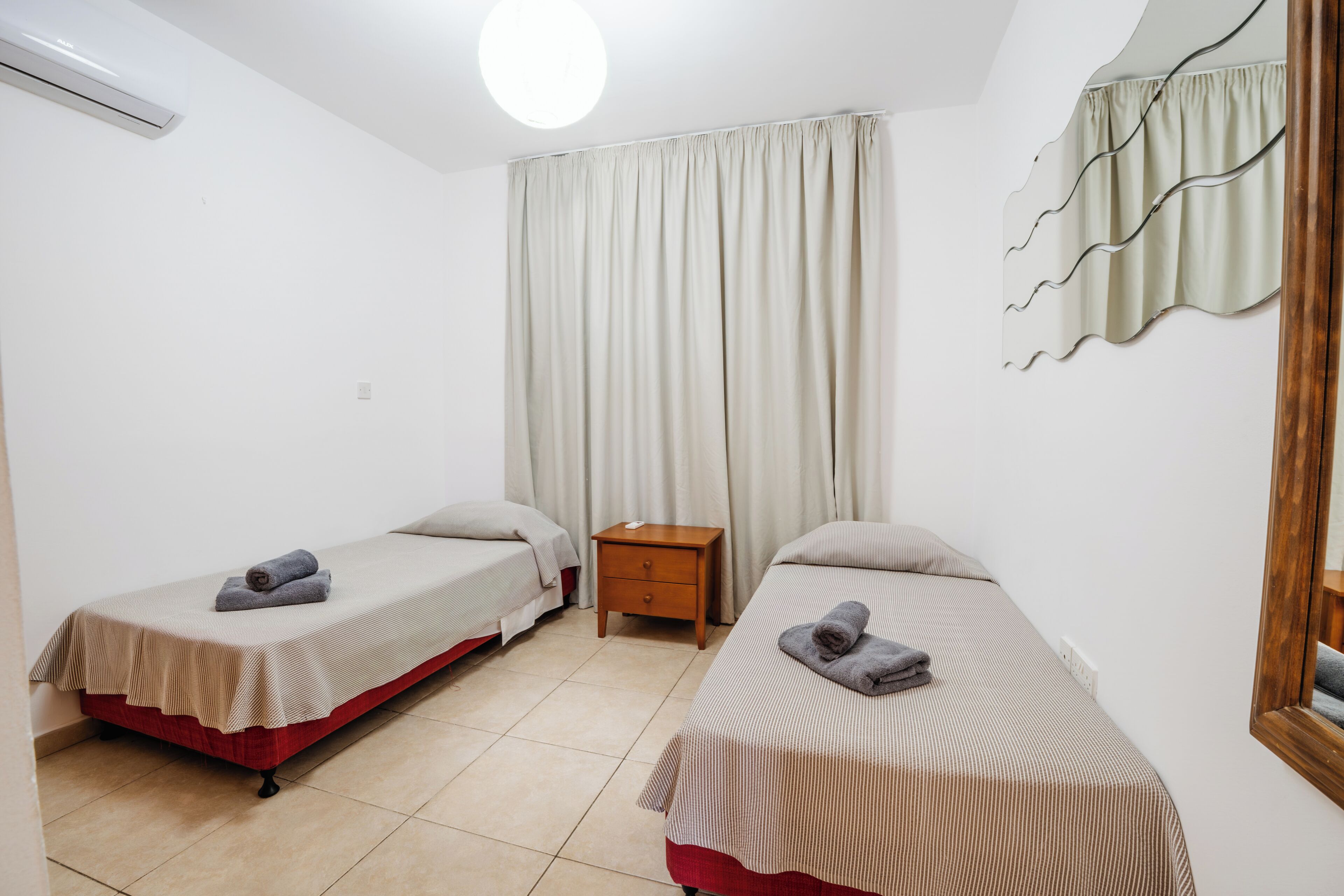 A room with two single beds, a wooden nightstand, a mirror, and a ceiling light.