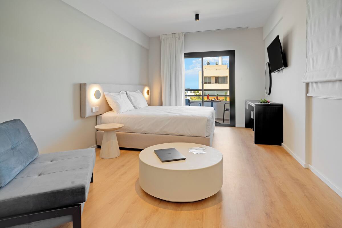 A modern hotel room with a bed, a round coffee table, a sofa, a TV, and a balcony with a view.