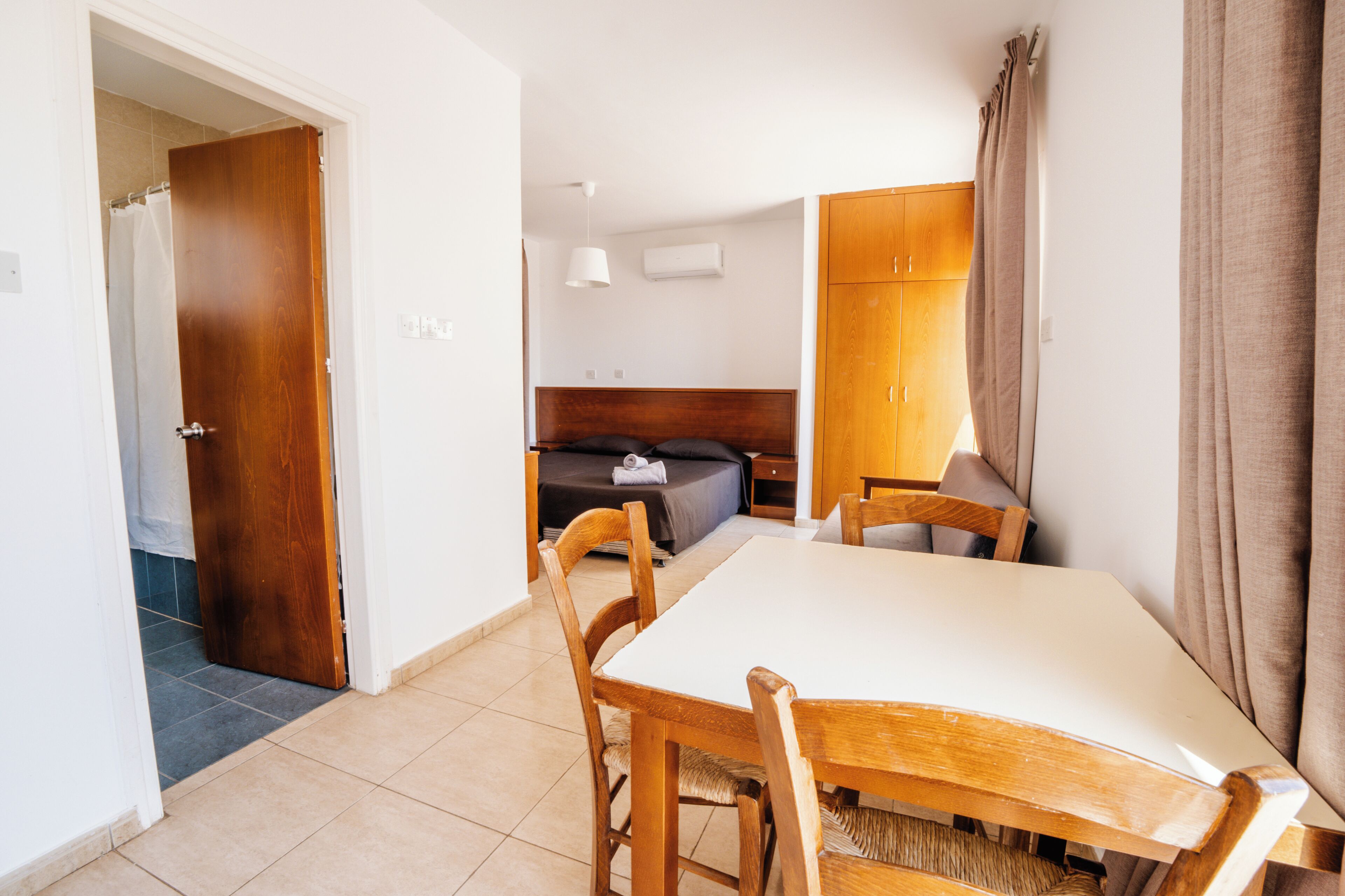 A hotel room with a wooden dining table, chairs, a bed with a headboard, a bedside table, and a wardrobe.