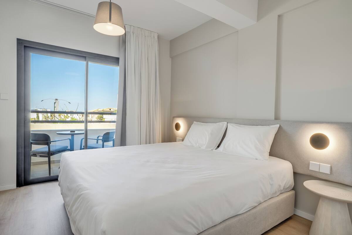 A modern hotel room with a large bed, a round wall light, a small table, and a view of the outside.