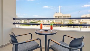 Un balcon avec deux chaises et une table, deux boissons, et une vue sur des immeubles et un ciel dégagé.