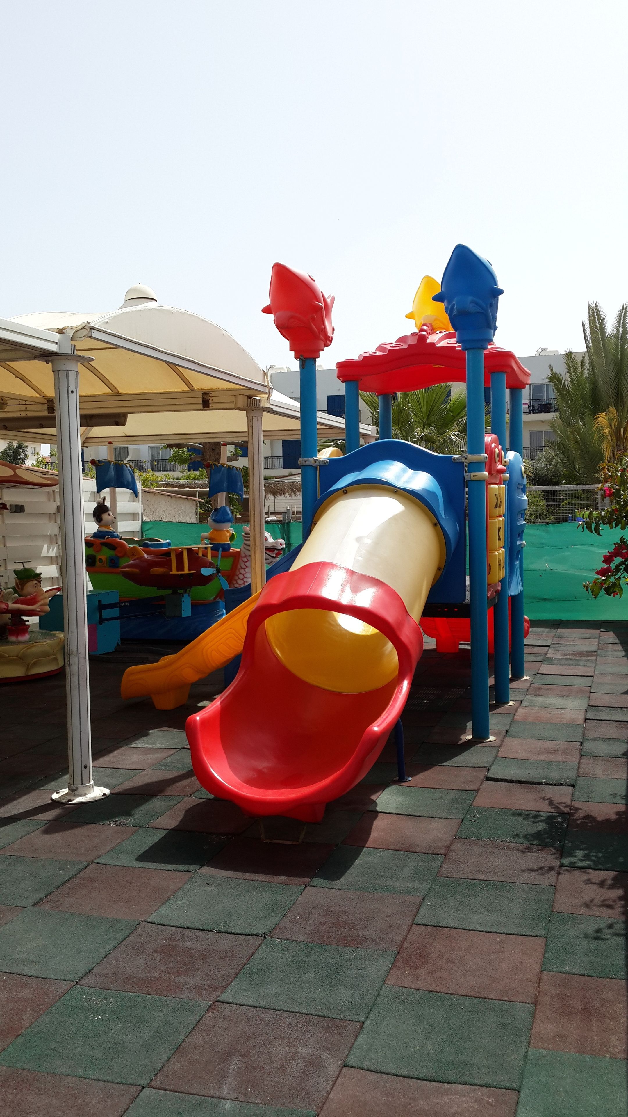 Un parque infantil con un tobogán rojo y amarillo, rodeado por una valla metálica y una zona cubierta.