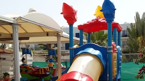 A playground with a red and yellow slide, surrounded by a metal fence and a covered area. - Captain Karas Collection (Paralimni)