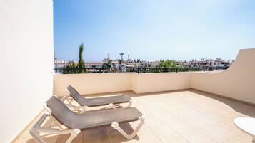 A rooftop terrace with two lounge chairs and a table, overlooking a cityscape with buildings and a clear sky.